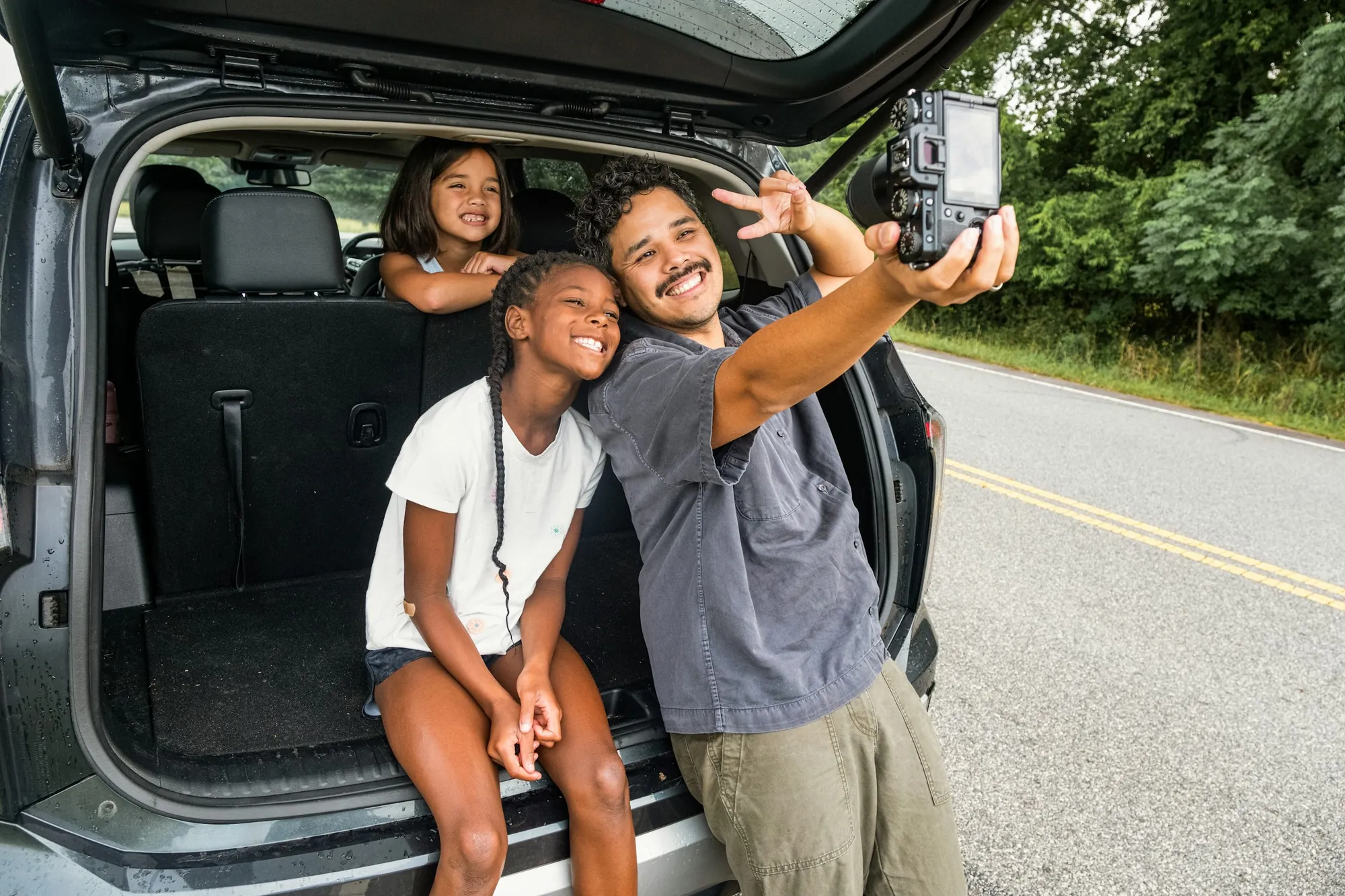 Pai e filhas sentados na bagageira do carro, a tirar uma fotografia numa estrada arborizada